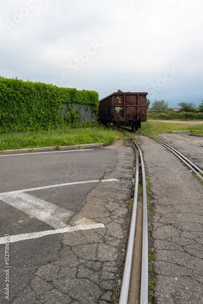 Fototapeta Rusty freight wagon on narrow rails disappearing into overgrown industrial area with cracked asphalt
