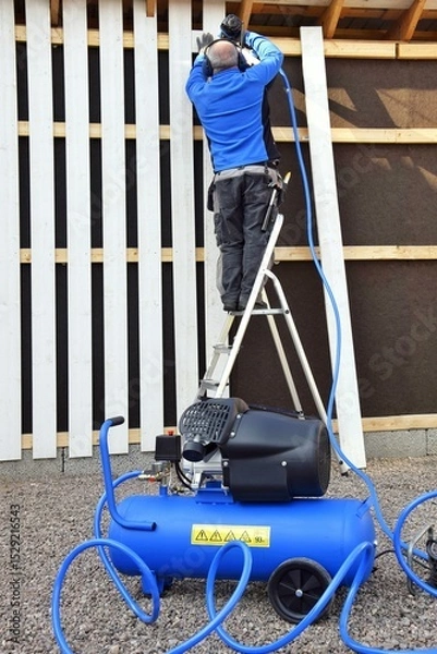 Obraz A man stands on a stepladder and holds a pneumatic stapler in his hand - a nail gun for a wooden board, hammering metal nails and staples using a compressor.