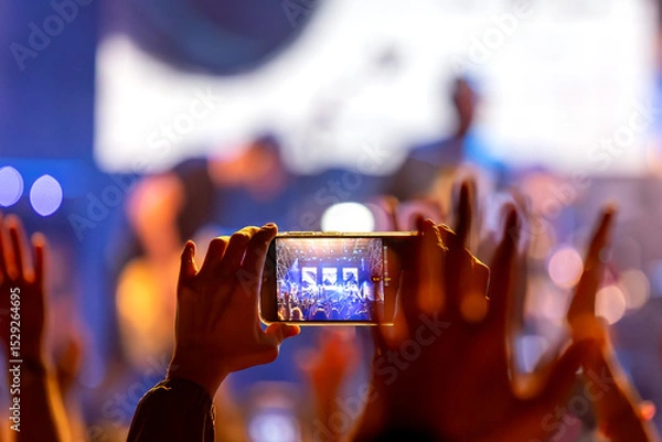 Fototapeta The audience records videos and takes photos with their smartphones during the concert of the musical band that performs on stage.