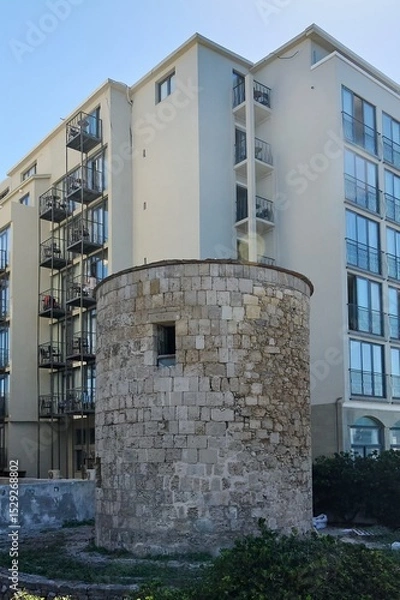Fototapeta An ancient stone tower stands prominently in front of a modern multi-story hotel building, showcasing the architectural blend of Rhodes, under a clear sky.