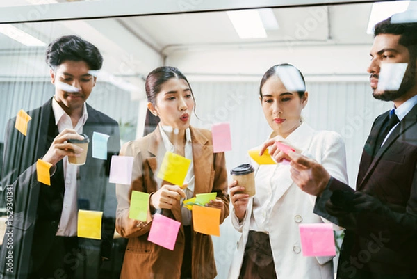 Fototapeta Multicultural office team collaborates in a brainstorming session, placing sticky notes on a glass wall. Business planning, creativity, and teamwork in a modern work environment with coffee in hand.