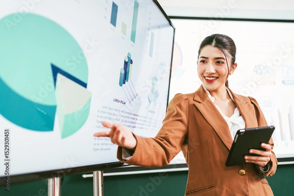 Fototapeta Confident businesswoman in a brown suit presenting financial charts on a large screen with a digital tablet in hand, showcasing analytic, teamwork, and strategic corporate planning in the office.