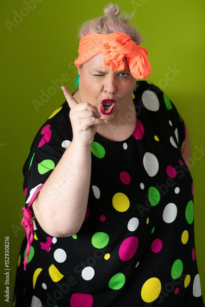 Fototapeta Plus size woman in vibrant polka dot outfit making a comic shrug gesture with outstretched hands and amusing expression.
