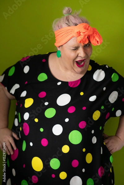 Fototapeta Plus size woman in vibrant polka dot outfit making a comic shrug gesture with outstretched hands and amusing expression.