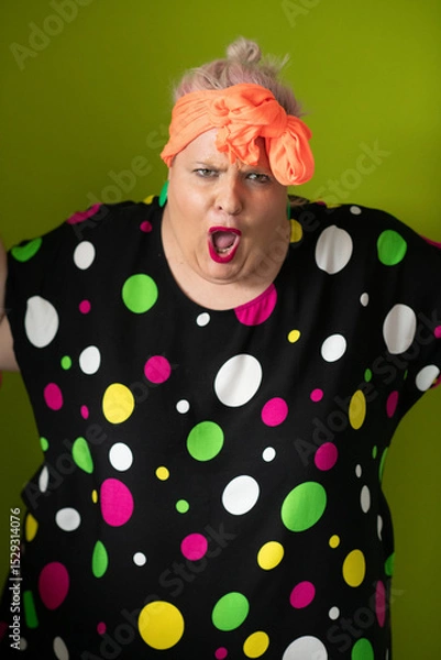 Fototapeta Plus size woman in vibrant polka dot outfit making a comic shrug gesture with outstretched hands and amusing expression.