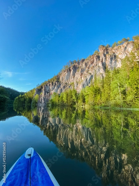 Obraz A vibrant and colorful kayak drifts peacefully across the tranquil river waters, while a magnificent mountain looms majestically in the background, forming a breathtaking natural scene