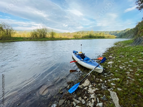 Obraz A brightcolored kayak rests peacefully on the sandy shore of a beautiful river, its vibrant hues contrasting nicely with the surrounding greenery and the shimmering water