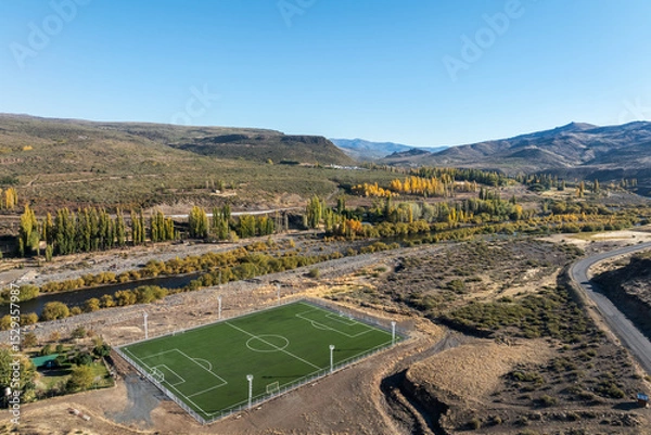 Fototapeta Aerial view of soccer field nestled in a valley with autumn foliage next to Nahueve river and distant hills. Villa del Nahueve, Neuquen, Argentina