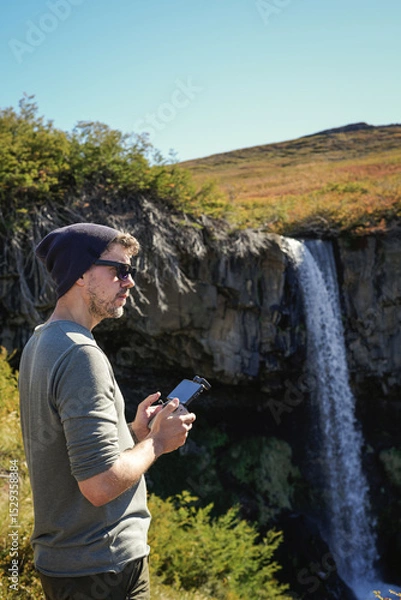 Fototapeta Man stands on a rock outcropping, holding drone controllernext to Cascada de la Usina waterfall. Caviahue, Neuquen, Argentina