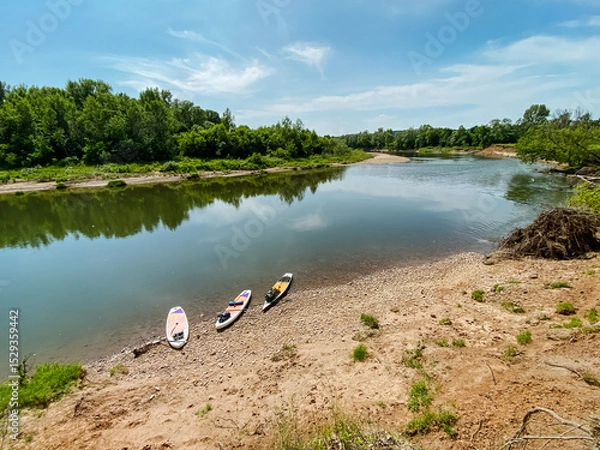 Obraz Three colorful kayaks are resting on the sandy shore of a beautiful river, waiting for adventurous paddlers to take them out onto the water for an exciting journey ahead