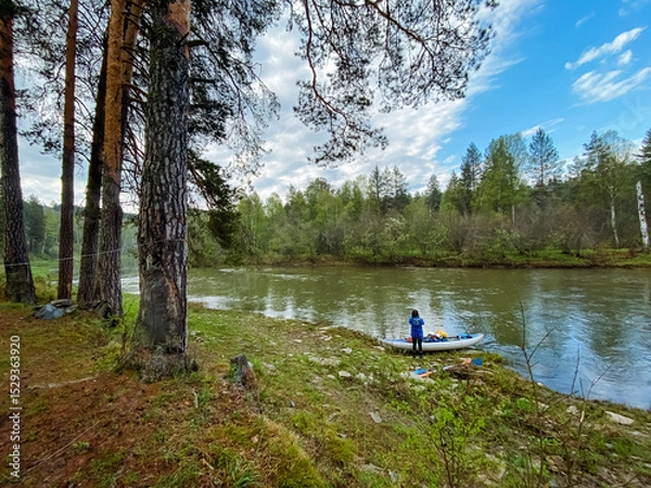 Obraz A person is sitting in a kayak on the sandy shore of a beautiful river, surrounded by lush trees and the stunning natural landscapes that define the wilderness and forested areas