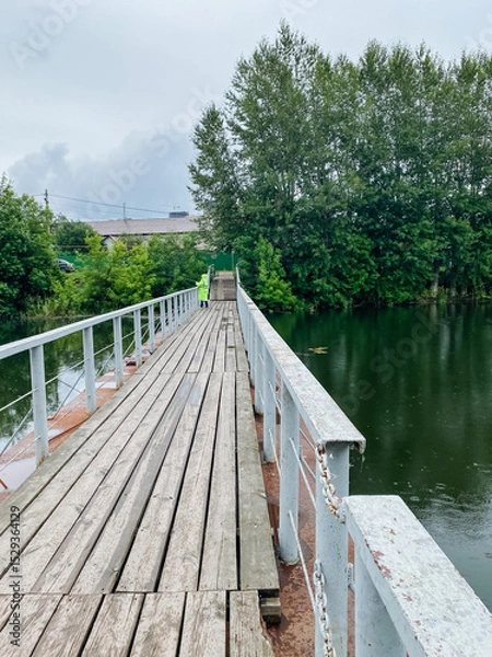 Obraz A charming wooden bridge gracefully spans over a tranquil body of water, surrounded by lush trees that provide shade and beauty to the scene, creating a picturesque landscape
