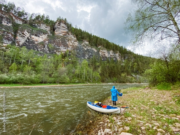 Obraz A man is standing on the peaceful shore of a river, right next to a sturdy raft that is moored nearby, enjoying the tranquil beauty of the natural environment and watercourse