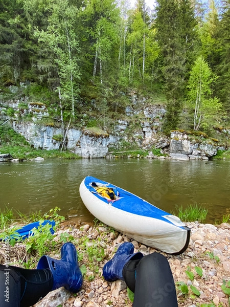 Obraz A person is comfortably seated next to a vibrant blue and white kayak that is resting on the sandy shore of a tranquil river, enjoying the peaceful beauty of nature all around them