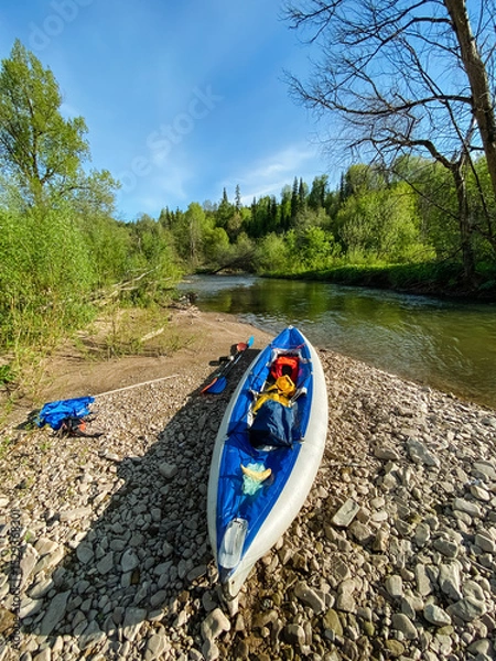 Obraz A vibrant blue and white kayak is currently resting on the rocky shore near a flowing river, creating a picturesque scene that captures the essence of outdoor adventure and recreation