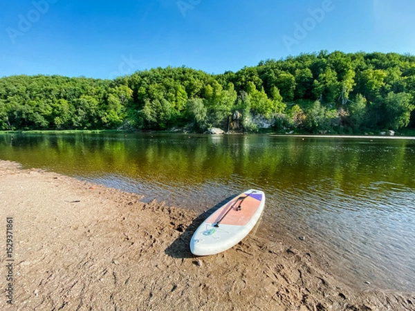 Obraz A colorful paddle board is currently resting on the warm sandy shore of a tranquil lake, waiting for someone to take it out onto the peaceful water surface for some fun and adventure