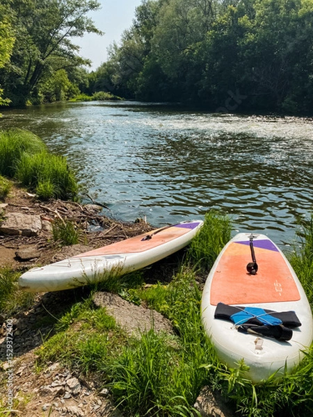 Obraz On a tranquil riverbank, two vibrant and colorful paddle boards are perfectly positioned, eagerly awaiting the fun activities and exhilarating water sports adventures that lie ahead