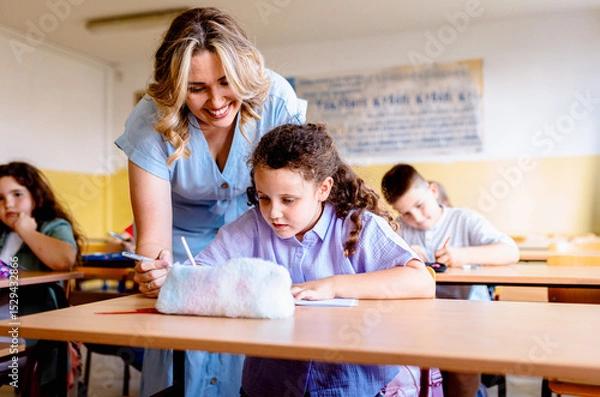 Obraz Smiling teacher assisting young girl with assignment at school desk in classroom.