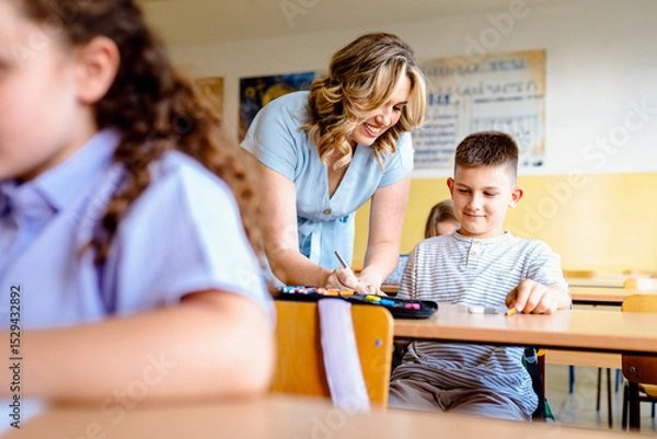 Fototapeta Smiling teacher assisting young boy with assignment at school desk in classroom.