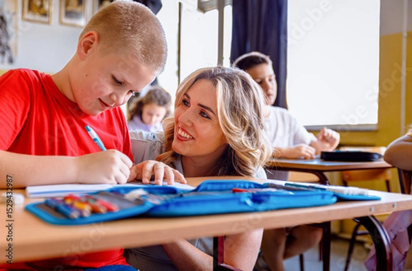 Obraz Smiling teacher assisting young boy with assignment at school desk in classroom.