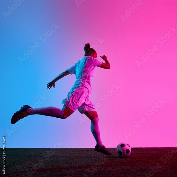 Fototapeta Female football player starting powerful kick toward ball during moment of action on field with neon gradient background. Concept of sport, impact, game, precision, motivation
