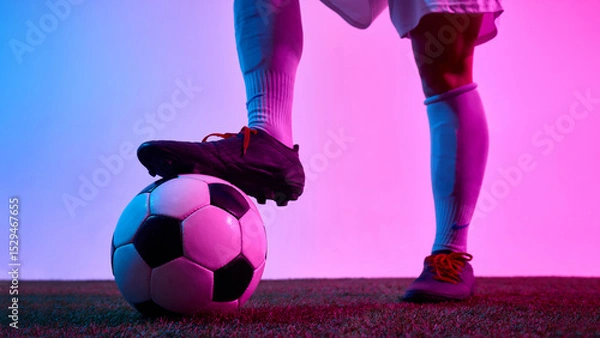 Fototapeta Detail view of female footballer's foot resting on ball on turf field under pink and blue lighting against gradient background. Concept of sport, control, readiness, visual power, brandimng.
