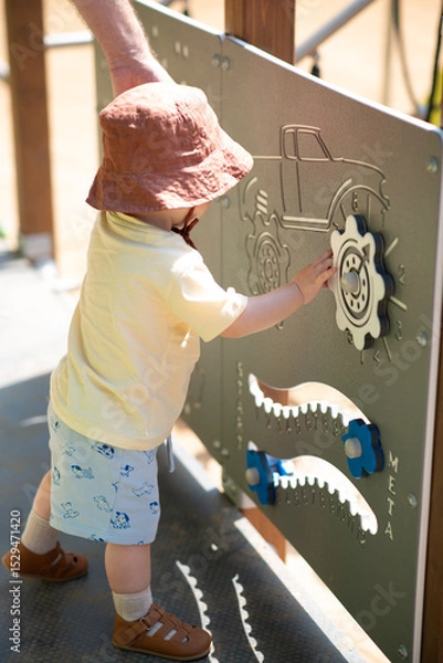 Obraz Baby boy dressed with summer cloves and hat is playing outside on the street, touching and exploring playground for little ones designed for sensory play. Lifestyle photo, sunny weather. 