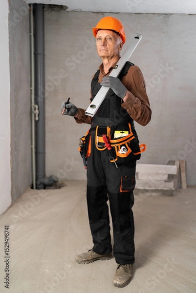 Obraz Elderly builder with tool belt holding wall measuring level and pencil looking at camera kindly. House construction and renovation