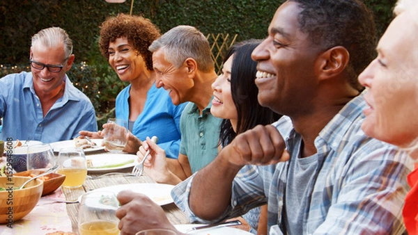 Obraz Group Of Mature Friends Sitting Around Table Enjoying Outdoor Meal In Backyard