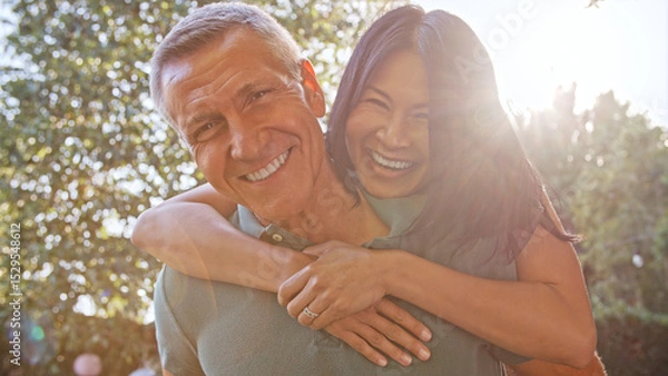 Obraz Outdoor Portrait Of Mature Couple With Woman Hugging Man From Behind With Lens Flare