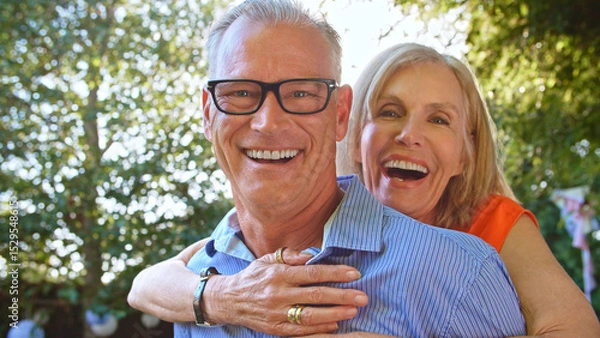 Obraz Outdoor Portrait Of Mature Couple With Woman Hugging Man From Behind With Lens Flare