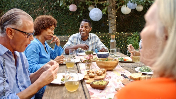 Obraz Group Of Mature Friends Sitting Around Table Enjoying Outdoor Meal In Backyard