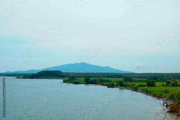 Fototapeta a calm sea view with agricultural rice fields and a backdrop of cloud-covered mountains.