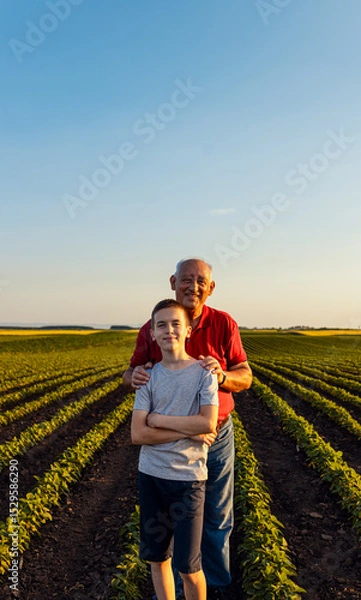 Obraz Portrait of senior farmer with his grandson standing in green soybean field and looking at camera.