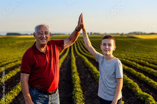 Obraz Portrait of senior farmer with his grandson standing in green soybean field and looking at camera.