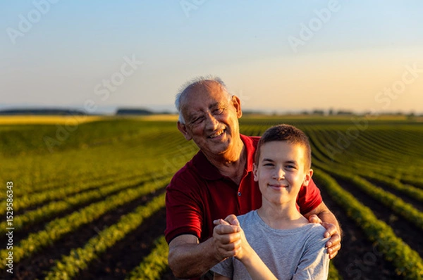 Obraz Portrait of senior farmer with his grandson standing in green soybean field and looking at camera.