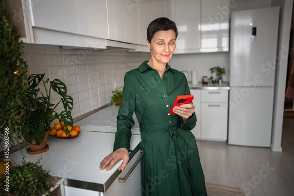 Fototapeta Laughing mature woman with smartphone on kitchen counter, absorbed in chat and social feeds. Digital connection, online interaction, tech dependency, retired modern habits, instant gratification