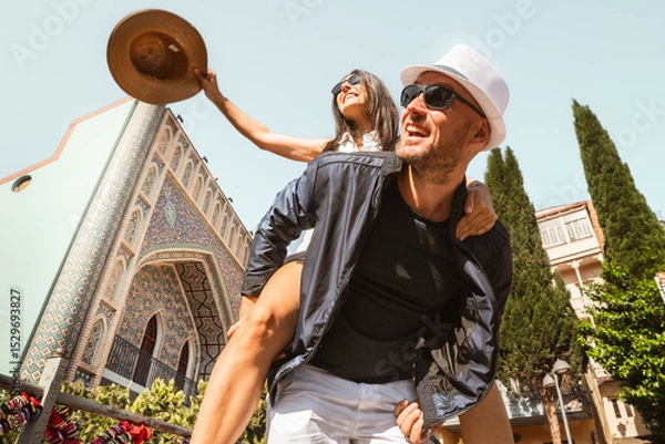 Fototapeta Happy couple enjoying sunny day, man carrying woman piggyback in front of colorful Persian-style mosaic building in Tbilisi old town. Tourist sightseeing famous Georgia attractions in capital Tbilisi