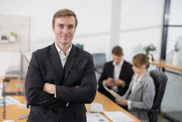 Fototapeta Caucasian businessman with arm crossed gesture in office while colleagues work on laptops in the background
