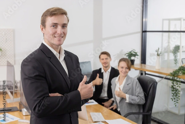 Fototapeta Caucasian businessman with thumb up gesture in office while colleagues work on laptops in the background