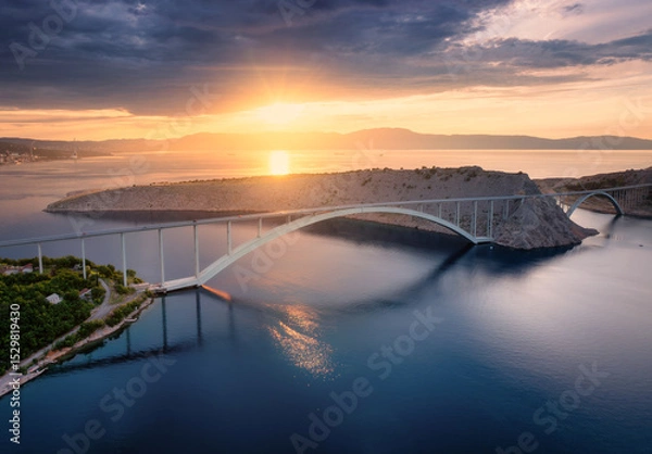 Obraz Aerial view of bridge and blue sea at colorful sunset. Modern Krk bridge island during golden hour, mountains, rocks, reflection in water and orange sky. Top drone view of bridges. Architecture