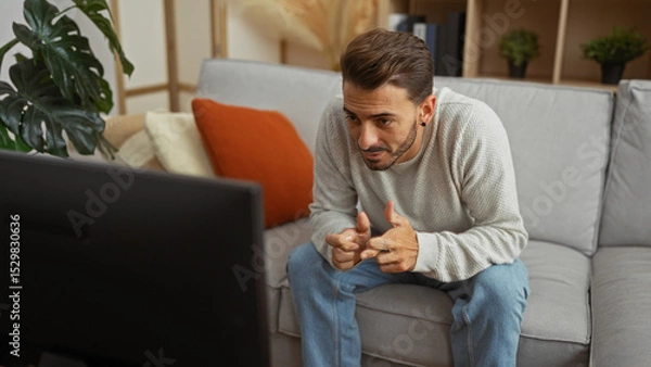 Fototapeta Young man relaxing in living room, watching television with focused expression, sitting on sofa, fingers interlocked, surrounded by cozy home decor, casual and stylish setting.
