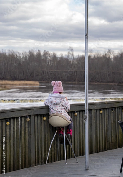 Fototapeta A child sits alone on a chair on a cafe terrace by the river.