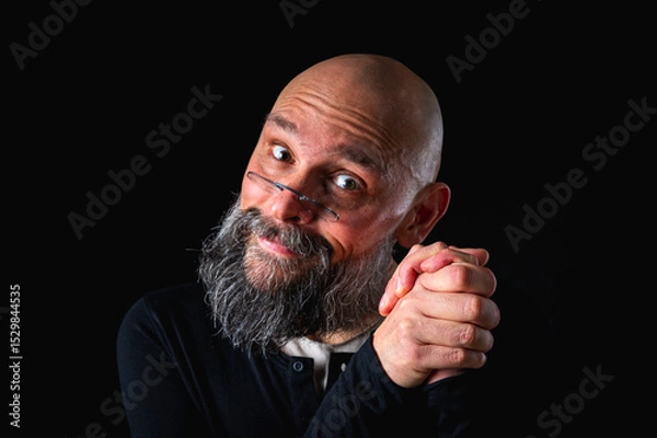 Obraz Portrait of a cunning-looking bald man with gray beard and glasses, clasping hands. Dark background. Mischievous or scheming expression. Concept of plotting, sarcasm, or ironic humor