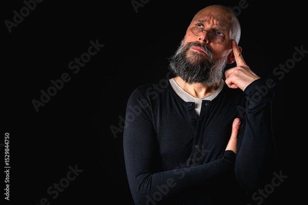 Obraz Thoughtful bald man with beard and glasses looking up, touching temple, deep in thought, isolated on black background. Concept of ideas, planning, decision-making