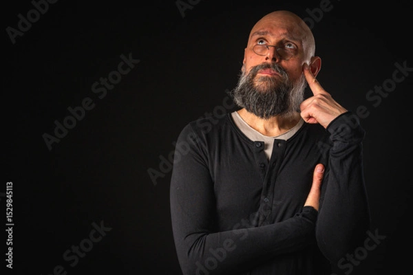 Obraz Thoughtful bald man with beard and glasses looking up, touching temple, deep in thought, isolated on black background. Concept of ideas, planning, decision-making