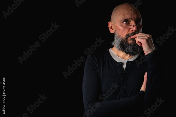 Obraz Thoughtful bald man with beard and glasses looking up, touching temple, deep in thought, isolated on black background. Concept of ideas, planning, decision-making