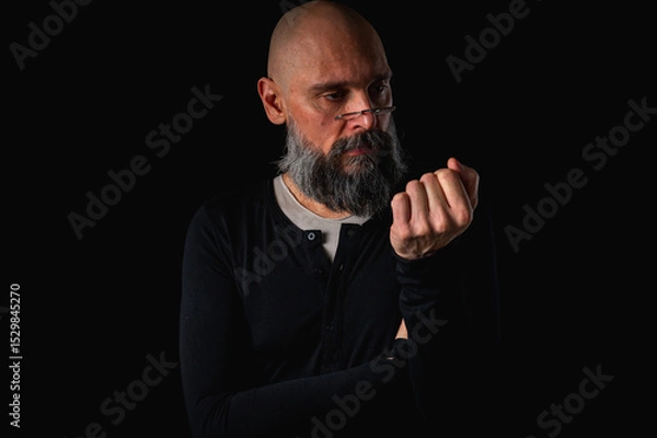Obraz Portrait of a cunning-looking bald man with gray beard and glasses, clasping hands. Dark background. Mischievous or scheming expression. Concept of plotting, sarcasm, or ironic humor