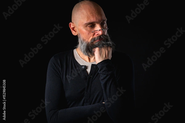 Obraz Thoughtful bald man with beard and glasses looking up, touching temple, deep in thought, isolated on black background. Concept of ideas, planning, decision-making