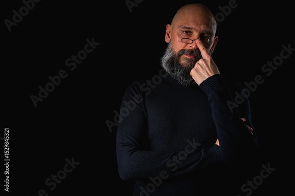 Obraz Thoughtful bald man with beard and glasses looking up, touching temple, deep in thought, isolated on black background. Concept of ideas, planning, decision-making